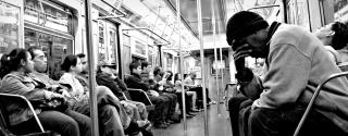 On a New York City subway (black and white photo), passengers sit. In the foreground, a man cradles his face in one hand. 