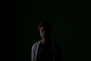 A Black woman looks at the camera, half-hidden in darkness