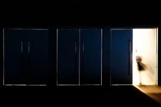 A series of doors in a dark room, with one door opening to allow in bright light as a blurred human figure moves through