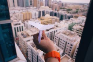 A hand holds a paper airplane in an open window, the tops of buildings visible underneath.