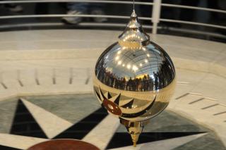 A gold Foucault's pendulum slowly swings across a marbled floor. 