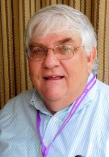 A portrait of Richard Nugent, an older white man. He is looking at the camera and has on glasses, a purple lanyard, and a blue striped shirt.