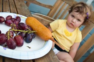 A toddler pouts in the background, while a plate of grapes with a corndog occupies the foreground.