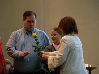 A minister handing a yellow rose to a church member, the lit chalice in the background