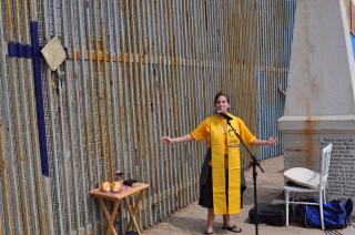 Minister in SSL stole stands at a mic next to Mexico border wall