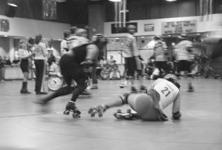 a blurred "action" shot, in black and white, of a roller participant falling on her hip while others roller-skate around her