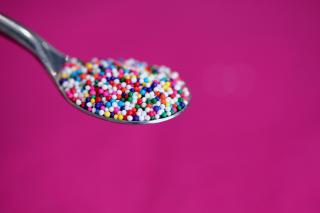 Against a fuschia background, a spoonful of tiny rainbow candy sprinkles