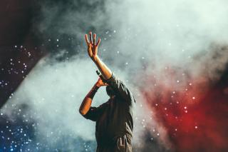 A singer with a microphone, reaching with dramatic body language, with smoke and lights in the background.
