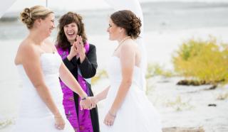 Two women in bridal gowns smile and their minister cheers as their wedding ceremony concludes.