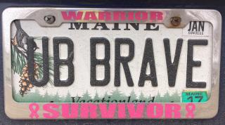 A Maine license plate that says "UB BRAVE." Its silver frame says "Warrior/Survivor," with pink breast cancer awareness ribbons.