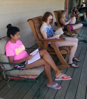 Three young women are reading books in chairs on a porch
