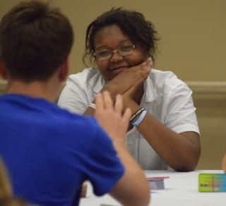 a young black woman wearing glasses looks encouragingly at a person who is facing her, speaking. that person is blurry and not the focus of the picture.