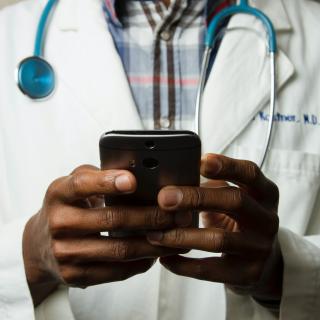 A person with dark skin, wearing a white doctor's coat and a stethoscope around their neck, holds a cell phone in their hands.