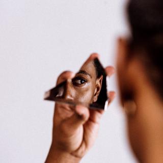 A black person holds a jagged piece of mirror, looking into it, as the camera captures the reflection oftheir eye.