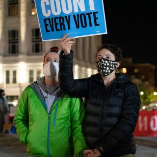 two people hold hands at a rally and display a sign reading, "Count Every Vote"