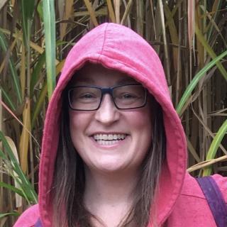 Laura Conkle smiles in an outdoor photo. They are wearing glasses and a pink hoodie.