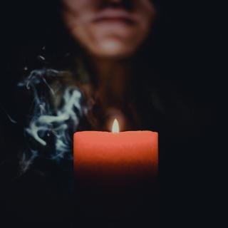 A red candle, lit and emanating a wisp of smoke, hovers in the dark. The lower half of a person's face is seen in the background.