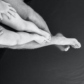 A black-and-white photo, taken from above, of the photographer's legs outstreched over water, and his young son nestled on top of his legs. 