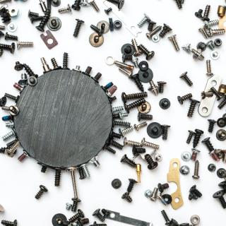 A scattering of hardware--nuts, bolts, screws, and washers--on a surface, and some of them clustering around a large circular magnet.