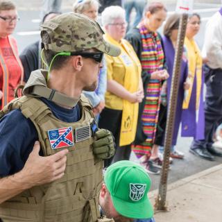 A white nationalist, wearing body armor with a white nationalist symbol, stands in front of a line of religious leaders, some wearing Side with Love garb, at the Charlottesville Unite the Right Rally (August 12, 2017).