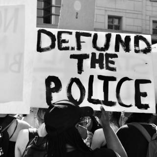 Black and white photo of a protester holding a sign that reads “DEFUND THE POLICE.” The protester is seen from behind and has dark skin and is wearing a baseball cap and a facemask.