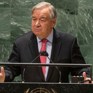 UN Secretary-General António Guterres is seen speaking at the podium of the UN General Assembly Hall. He is wearing a black suit with a red tie.