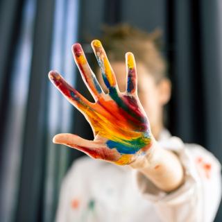 A person holds their palm to the camera, obscuring their face. Their palm is painted with streaks of bright paint in yellows, reds, and blues.