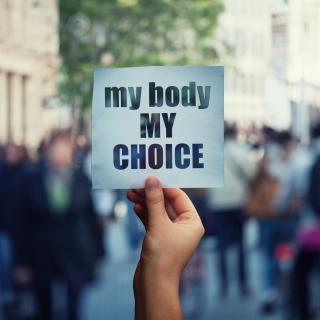 A white person's hand holding a sign with the message "my body my choice" over a crowded street