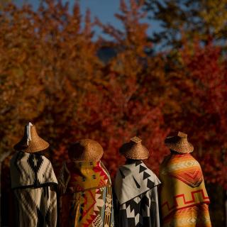 Four members of the Squamish and Lil'wat Nations, with their backs to the camrea, are each draped in a traditional Squamish or Lil'wat blanket. 