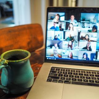 A ceramic mug of coffee sits on a table next to an open laptop, showing many people in Zoom cells.