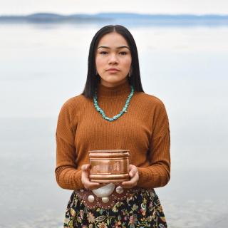 Photo of teenage water protector Autumn Peltier standing in front of a lake under an overcast grey sky. She is holding a copper-colored vessel.