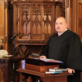 Andrew speaks from a pulpit, wearing a black clergy robe.