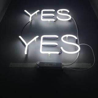 Against a dark background, a neon message in silver reads YES YES