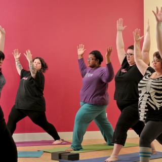 In a yoga studio, called Fat Yoga, seven people move into a powerful pose on their yoga mats.
