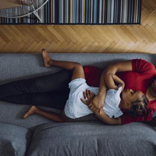 A photo, from above, of a Black mother with daughter lying down on cozy couch in living room, cuddling and napping together at home.
