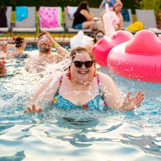 A fat woman in a bikini laughs, in a swimming pool, surrounded by other fat and laughing people and their pool toys.