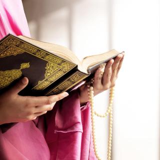 A person in pink robes, mostly cropped out of the photo, holds a Qur'am and Muslim prayer beads.
