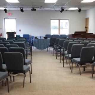 Inside a small congregation sanctuary, with about 50 chairs in foreground and a podium, table and piano in the background