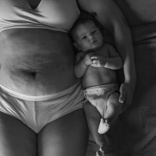 A newborn infant lies on a bed next to their mother, whose torso reveals stretch marks and other signs of recently giving birth.