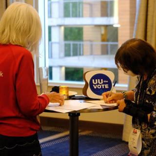 Two people stand at a table that has a UU the Vote sign on it, writing