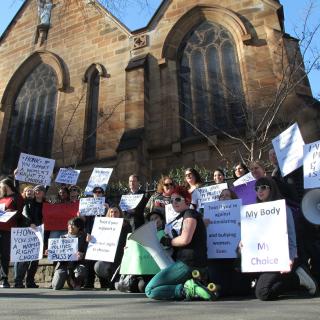 In front of a gothic church, an assembly of people holding up pro-choice signs to traffic.