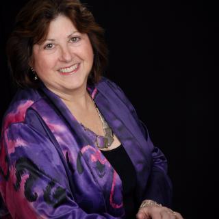 In a studio portrait setting, Deb smiles broadly with her hands crossed in front of her. She's wearing a purple silk jacket. 