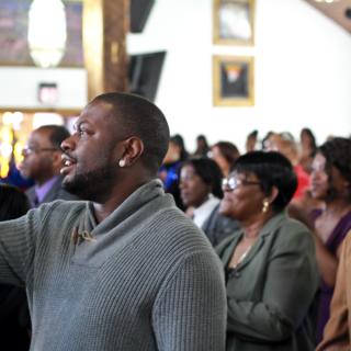 Inside a church, Black people stand as if listening to a preacher or choir.