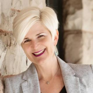 Heather McDuffee smiles broadly. She is a white woman with a short platinum blonde hair cut, red lipstick, and a blazer.