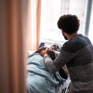 A Black person with their back to the camera sits, holding the hand of a Black elder who's lying in a hospital bed.