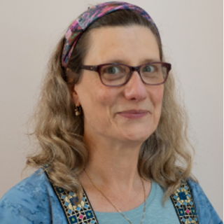 Headshot of Rev. Ellen Spero, she is a light skinned person with shoulder-length hair, wearing a blue blouse, purple headband, and black-rimmed glasses.
