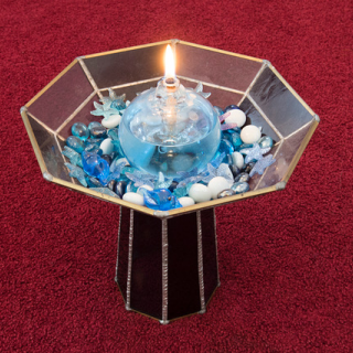 A lit glass globe sits in a bed of stones and gems atop a dark glass chalice. The chalice is on maroon carpet.