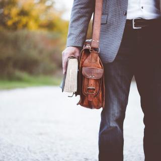 Torso and upper legs of a person in jeans and a tweed jacket carrying a thick book and leather messenger bag.