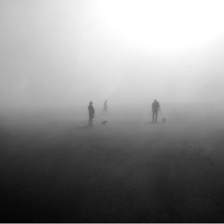 Three people and two dogs wander in the distance in heavy fog. The image is in black and white.