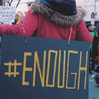 At a March For Our Lives event, a protester is seen from behind wearing a very large sign that says #Enough